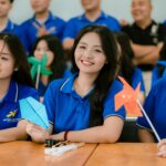 Home Group of cheerful students in blue shirts holding colorful pinwheels indoors.